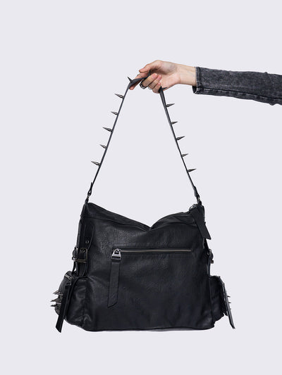 Black leather handbag with silver spikes on the strap, held by a hand, featuring buckle details and a rugged, edgy design.