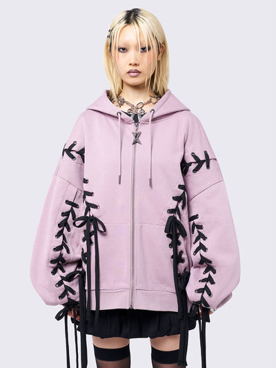 Pink zip-up hoodie with black lace corset details along the sleeves and front. Features a drawstring hood, zip fastening, long sleeves, and an oversized fit.