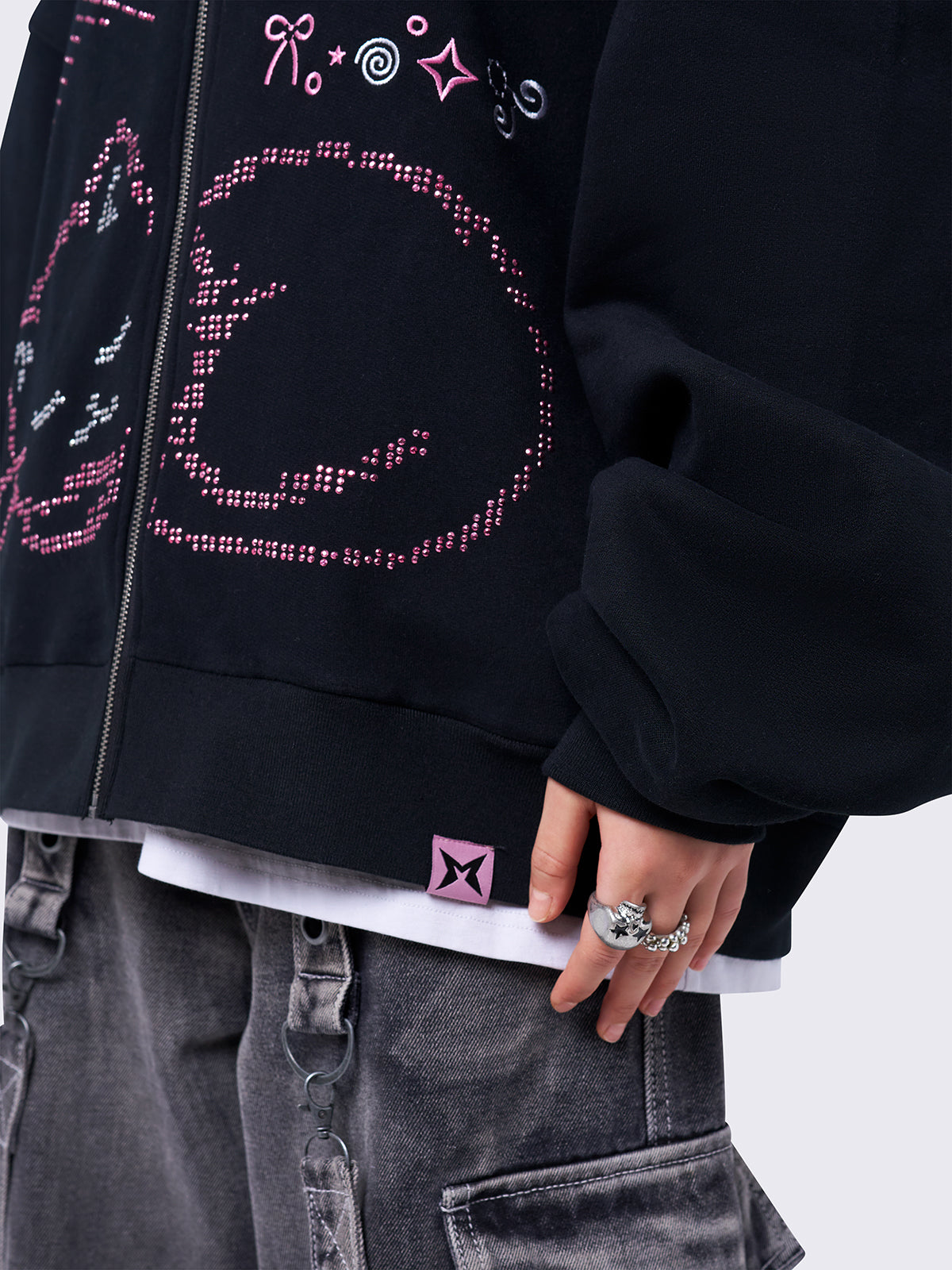 Black zip-up hoodie jacket featuring a pink cat and stars graphic with stud and embroidery details. Designed with a drawstring hood, zip fastening, long sleeves, ribbed trims, and an oversized fit.