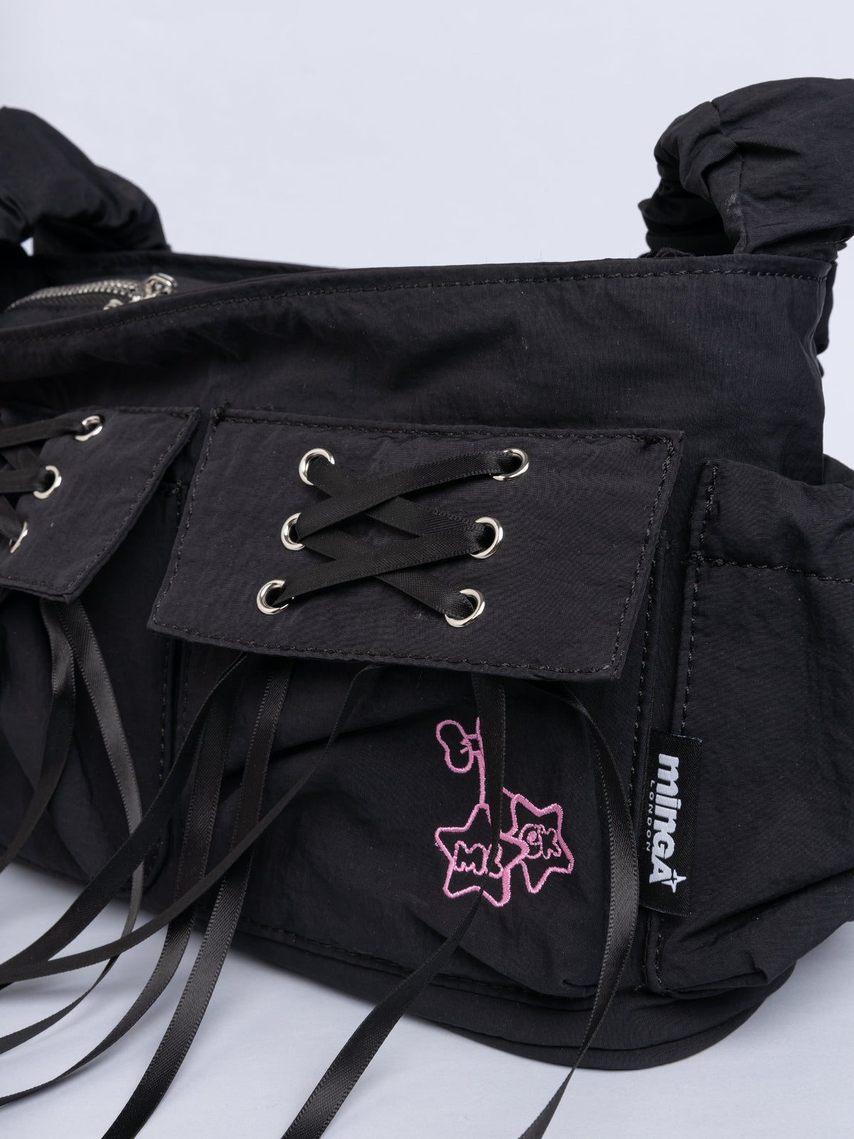 Black shoulder bag with ruched strap, lace-up ribbon front pockets, detachable plush pink bunny charm, and multiple pockets.