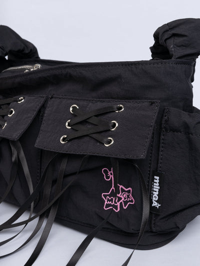 Black shoulder bag with ruched strap, lace-up ribbon front pockets, detachable plush pink bunny charm, and multiple pockets.