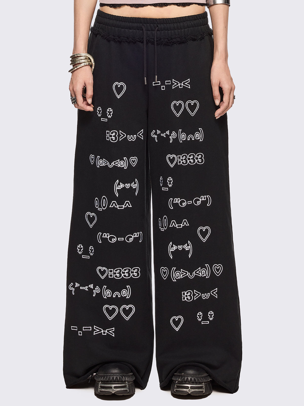 Black oversized sweatpants with all-over white emoticon and heart graphics in a playful ASCII/kaomoji style, styled in a relaxed streetwear fit.