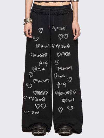 Black oversized sweatpants with all-over white emoticon and heart graphics in a playful ASCII/kaomoji style, styled in a relaxed streetwear fit.