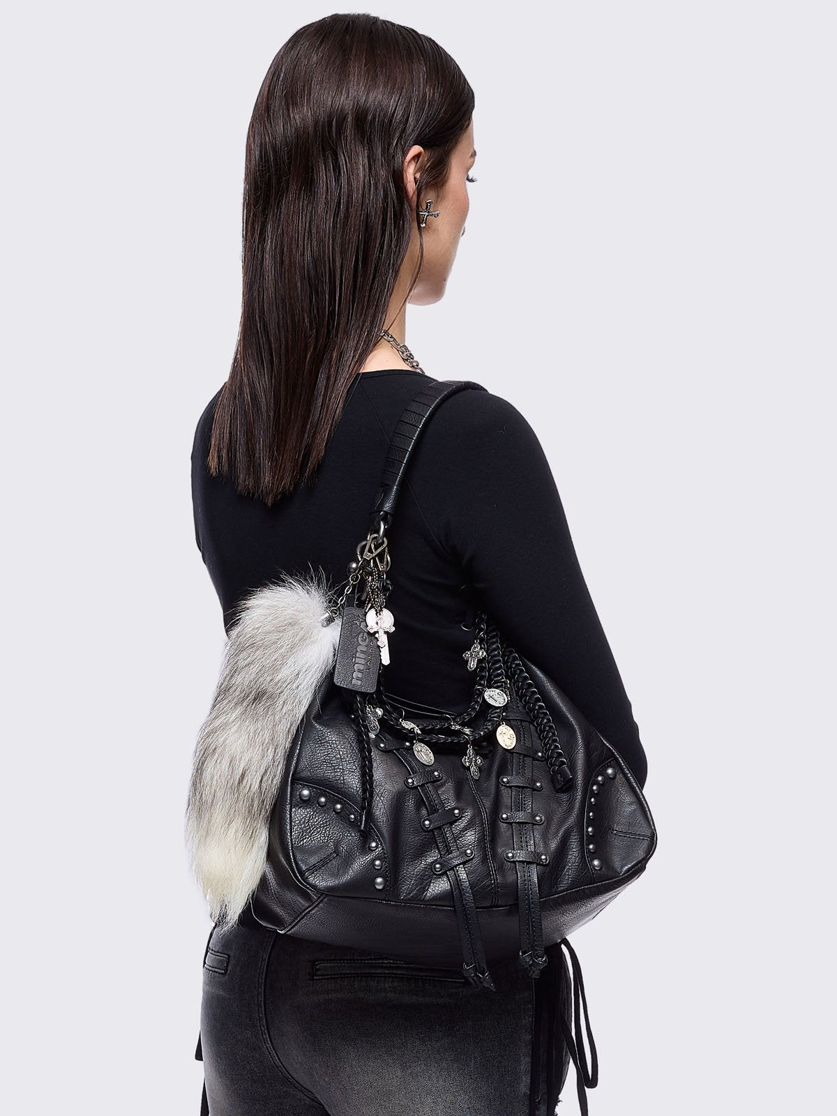 A black leather hobo bag decorated with silver studs, metallic charms, braided leather, and a large grey and white fur tail charm.