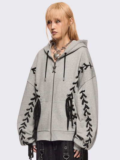 Grey marl zip-up hoodie with black lace corset details along the sleeves and front. Features a drawstring hood, zip fastening, long sleeves, and an oversized fit.
