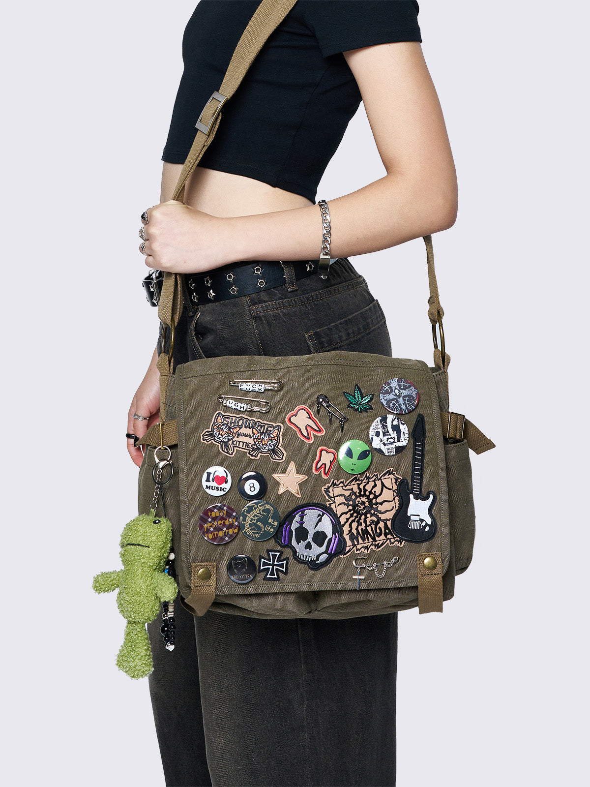 Olive green canvas messenger bag with grunge-inspired patches and pins, featuring a guitar, skull, alien, and other edgy designs. Includes a frog plush keychain and adjustable strap for a Y2K, alternative look.