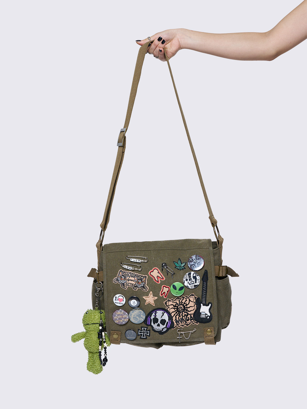 Olive green canvas messenger bag with grunge-inspired patches and pins, featuring a guitar, skull, alien, and other edgy designs. Includes a frog plush keychain and adjustable strap for a Y2K, alternative look.