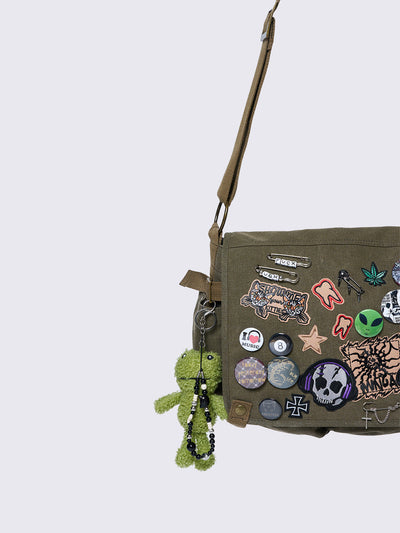 Olive green canvas messenger bag with grunge-inspired patches and pins, featuring a guitar, skull, alien, and other edgy designs. Includes a frog plush keychain and adjustable strap for a Y2K, alternative look.