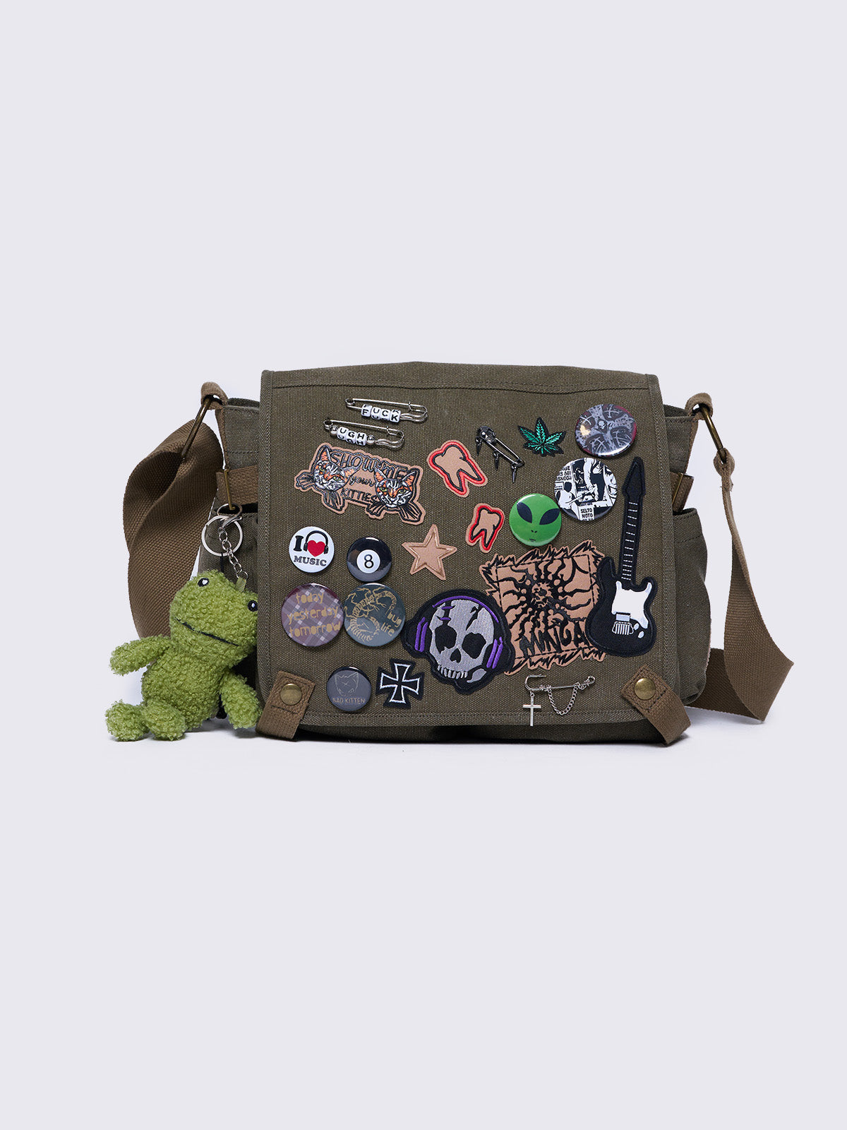 Olive green canvas messenger bag with grunge-inspired patches and pins, featuring a guitar, skull, alien, and other edgy designs. Includes a frog plush keychain and adjustable strap for a Y2K, alternative look.