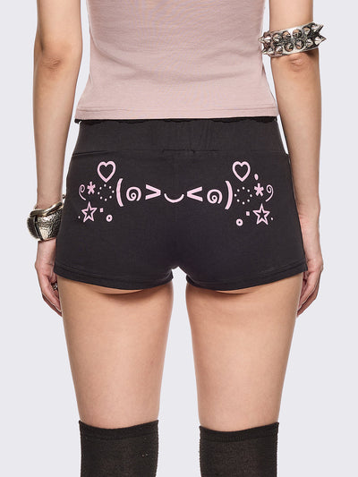 Black sweat micro shorts featuring kaomoji ASCII-style graphic print on side and back and a front pink bow detail. Low rise and elasticized waist.